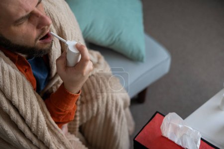 A sick man wrapped in a blanket treats a cold at home using a spray for a sore throat. High angle view