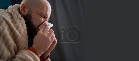 Braucht Behandlung und Medikamente. Niesen, laufende Nase, Husten. Kranker Mann sitzt zu Hause auf dem Sofa und niest in Serviette. Banner. Kopierraum