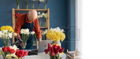 Ein grauhaariger Mann mittleren Alters, Florist, arbeitet selbstbewusst mit einem Laptop in einem Blumenladen. In der einen Hand hält er einen Strauß Narzissen
