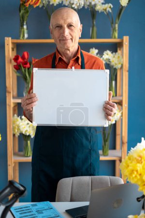 Un anciano sonriente de piel clara vendedor de flores tiene una pizarra blanca en sus manos. Detrás de él hay estantes con flores..