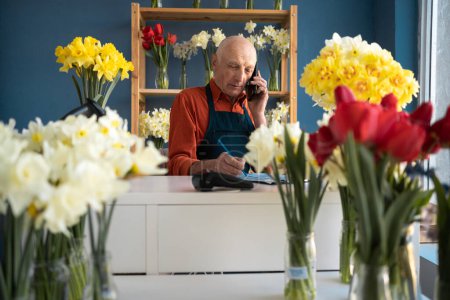 Feliz anciano vendedor de flores hablando por teléfono y escribiendo la entrega de un ramo con un lápiz. Hay muchos ramos hermosos a su alrededor.