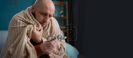Ein bärtiger müder Mann, der sich krank fühlt und Tabletten in der Hand hält, sitzt auf dem Sofa in seinem Wohnzimmer. Banner. Kopierraum