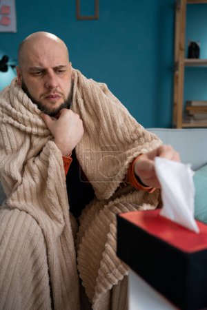Téléchargez les photos : Malade attrayant homme ont une fièvre, la grippe et prendre papier mouchoirs éternuement nez, écoulement tout en étant assis sur le canapé. Gros plan - en image libre de droit