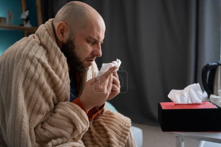 Sick man suffering from running stuffy nose and sore throat. Upset ill guy blowing her nose using paper napkin tissue sitting on sofa, Side view