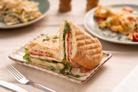 Toasted panini with ham, cheese and lettuce sandwich on plate serving on table with another dishes for breakfast. Copy space