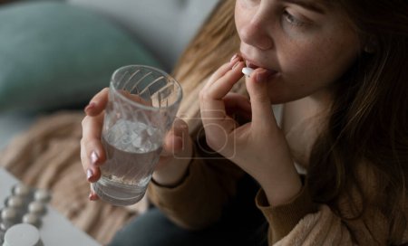 Jeune femme caucasienne malsaine assise sur un canapé dans le salon, couverte d'une couverture, tenant une pilule et un verre d'eau.