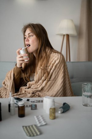 a young beautiful Caucasian sick girl sits at home on the sofa, wrapped in a blanket, uses a cough spray for the throat. The concept of treating viruses for a healthy lifestyle in the cold season