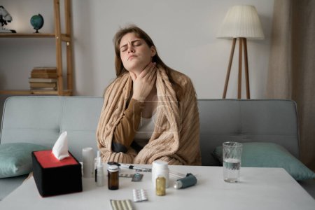 Halsschmerzen. Hände einer kaukasischen Frau, die ihren wunden Hals berührt. Sie leidet unter Halsschmerzen und Schluckbeschwerden. Zuhause auf dem Sofa sitzend, in eine Decke gehüllt. Kaltes Konzept.