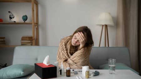 Eine junge kranke Frau sitzt zu Hause auf dem Sofa, beugt sich über einen Tisch mit Tabletten und hält ihren Kopf mit der Hand. Erkältungs- und Kopfschmerzkonzept.
