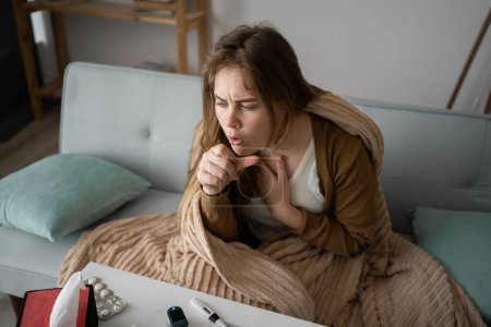 view from above. young sick upset woman sitting on the sofa at home covered with a blanket coughing intensely, sick girl with flu symptoms. copy space. banner