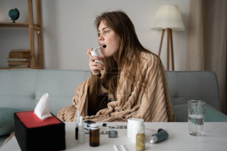 a young beautiful Caucasian sick girl sits at home on the sofa, wrapped in a blanket, uses a cough spray for the throat. The concept of treating viruses for a healthy lifestyle in the cold season