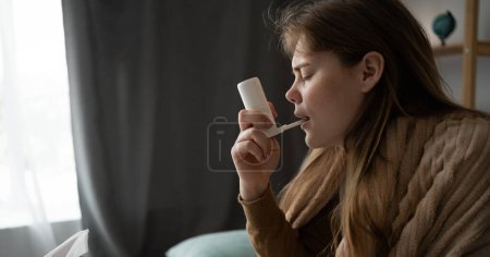 side view of a sick girl sitting at home on the sofa, wrapped in a blanket, using a cough spray for her throat. Healthy lifestyle virus treatment concept. Banner. copy space.