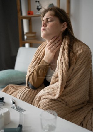Halsschmerzen. Hände einer kaukasischen Frau, die ihren wunden Hals berührt. Sie leidet unter Halsschmerzen und Schluckbeschwerden. Zuhause auf dem Sofa sitzend, in eine Decke gehüllt. Kaltes Konzept.