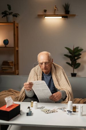 elderly sick man, wrapped in blanket, reads instructions for taking pills. Disease. Treatment. copy space