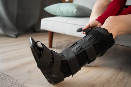 woman in orthosis sits on sofa after car accident. Attaching orthosis or bandage to support the ankle joint after leg surgery. Close-up. Copy space