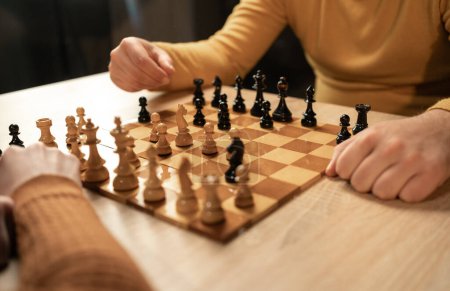 Close-up view of two men playing chess at home. The focus is on their hands and the wooden chessboard as they make strategic moves in an indoor setting. Hobby and leisure concept