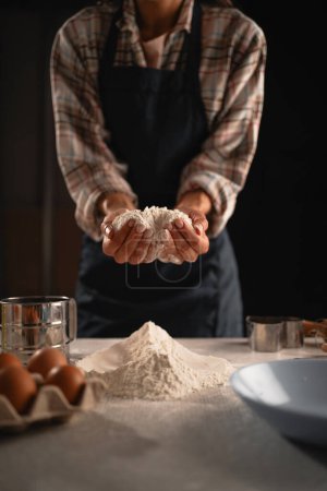 A chef in a plaid shirt and apron handling flour over a kitchen counter with eggs and kitchen utensils.