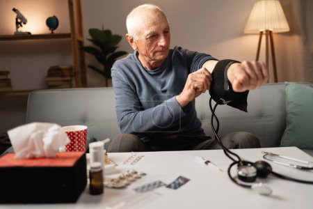Ein älterer Mann sitzt auf einer Couch und misst mit einem Blutdruckmessgerät seinen Blutdruck. Verschiedene Medikamente liegen auf dem Tisch und unterstreichen den Fokus auf Gesundheit und Selbstkontrolle zu Hause..
