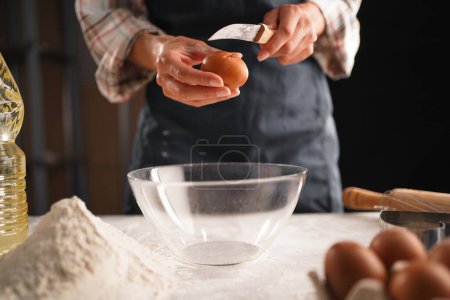 Close-up of female hands cracking an egg with a knife over a clear bowl. Flour, eggs, and a bottle of cooking oil are on the table. Preparing dough for baking in a home kitchen.