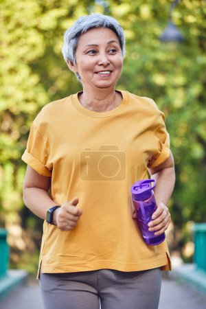 Téléchargez les photos : Mature fit femme asiatique en tenue de sport tenant bouteille d'eau faire de l'entraînement à l'extérieur, le jogging dans le parc d'été. Mode de vie sain et actif des retraités, concept de perte de poids - en image libre de droit
