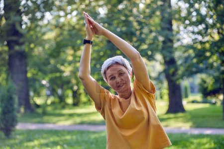 Photo for Older active Asian woman in sportswear look at camera meditating in summer park with folded raised palms makes stretching exercises, do yoga work out practice outdoor. Healthy lifestyle on retirement - Royalty Free Image