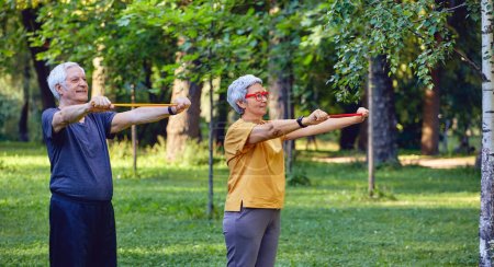 Photo for Senior 60 spouses wear sportswear doing hands exercises, training arms outdoor in summer park in the morning using resistance rubber bands. Healthy lifestyle, active retired life use modern appliance - Royalty Free Image