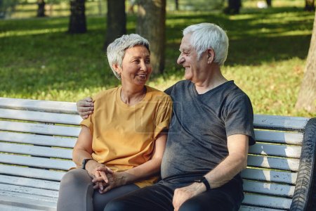 Téléchargez les photos : Heureux couple multi racial de cheveux gris âgés reposant sur un banc dans le parc après une promenade, promenade matinale souriant profiter de la détente en plein air à la journée ensoleillée d'été. Fit famille, pause, concept d'amour - en image libre de droit