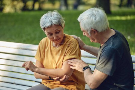 Photo for During morning sportive stroll or making exercises in park, old 60s woman got injured her shoulder or elbow gripping arm sit in bench with caring disappointed husband. Traumas of older people concept - Royalty Free Image
