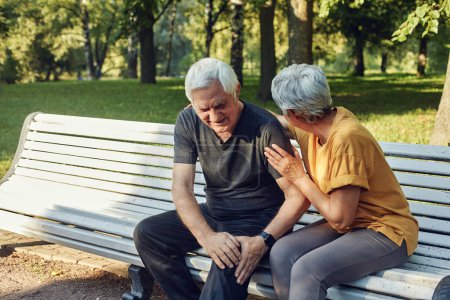 Téléchargez les photos : During morning sportive stroll or making exercises in a park, elderly 60s man got injured his knee, gripping leg sit in bench with caring disappointed wife. Traumas, injures of older people concept - en image libre de droit