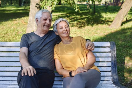Téléchargez les photos : Heureux couple multi racial de cheveux gris âgés reposant sur un banc dans le parc après une promenade, promenade matinale souriant profiter de la détente en plein air à la journée ensoleillée d'été. Fit famille, pause, concept d'amour - en image libre de droit