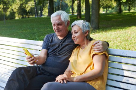 Photo for Mature spouses sit on bench in summer park with smart phone. Grey-haired couple look at cellphone screen enjoy new application outdoor use modern tech, enjoy online fun together spend time outside - Royalty Free Image