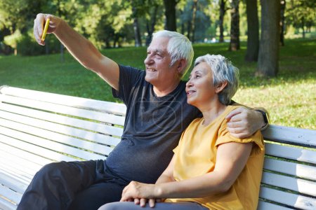 Téléchargez les photos : Heureux conjoints aînés prennent selfie photo assis sur le banc dans le parc d'été. Couple aux cheveux gris regarder l'écran de téléphone portable faire vidéocall utilisation extérieure technologie moderne, profiter du plaisir en ligne ensemble passer du temps à l'extérieur - en image libre de droit