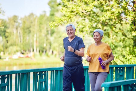 Téléchargez les photos : Femme asiatique et caucasienne d'âge moyen et mari portent des vêtements de sport se promenant le long du pont dans le parc d'été, profiter de la promenade sportive matinale ensemble à l'extérieur. Mode de vie sain et retraite - en image libre de droit