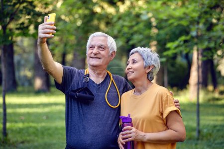Photo for Old sportive couple staring at smart phone cam take selfie picture enjoy modern wireless tech usage while stroll in sportswear in summer park. Carefree retired life, lifestyle using technology concept - Royalty Free Image