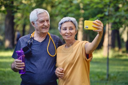Photo for Old sportive couple staring at smart phone cam take selfie picture enjoy modern wireless tech usage while stroll in sportswear in summer park. Carefree retired life, lifestyle using technology concept - Royalty Free Image