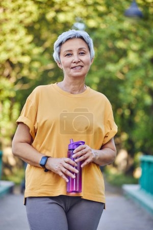 Photo for Older woman in sportive clothes holds reusable plastic bottle standing in footpath in summer park, caring about health, relieving thirst during morning work out. Healthy lifestyle - Royalty Free Image