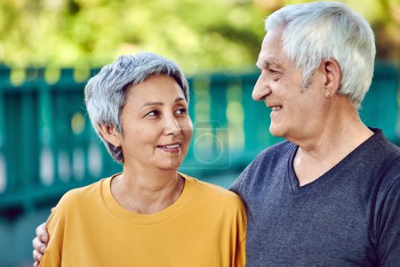 Foto de Optimistic attractive multiracial elderly 60s sporty couple pose in park smiling looking at each other with tenderness and love. Happy endless marriage, romantic relations, family concept - Imagen libre de derechos