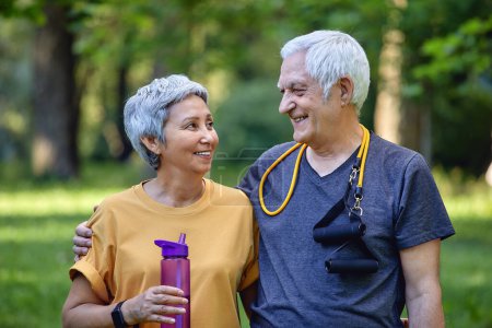 Photo for Attractive older couple in sportswear hugging pose look at each other standing in summer park after morning stroll, sport active lifestyle holds bottle with still water. Healthy retired life - Royalty Free Image