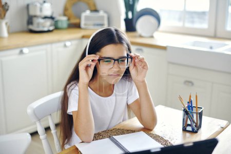 Téléchargez les photos : Jolie fille 12s porter des écouteurs et des lunettes font tâche, étudier en ligne, éprouve des problèmes de vision, plissant, ayant des chutes de vue, des problèmes de santé assis à table dans la cuisine. Mauvaise vision, technologie moderne - en image libre de droit