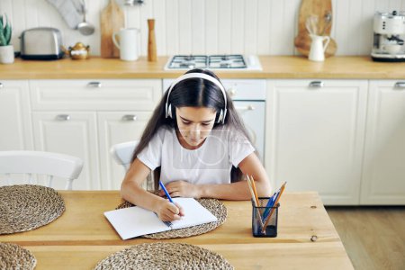 Téléchargez les photos : Jolie fille pré-adolescente dans la tâche d'écriture casque sans fil, rend l'affectation s'asseoir à table dans la cuisine, semble concentré, engagé dans l'exercice. Enseignement à domicile seul à la maison - en image libre de droit