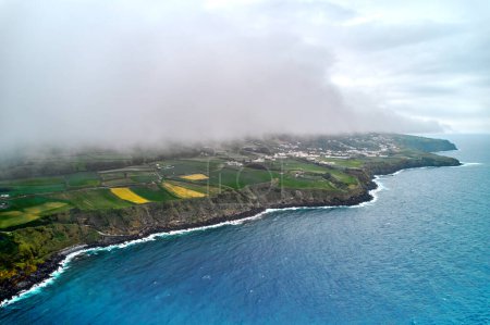 Photo for Aerial shot, drone point of view of Atlantic ocean waters, rocky shoreline, misty clouds over agricultural fields of Ponta Delgada Island during cloudy weather. Sao Miguel, Azores, Portugal - Royalty Free Image