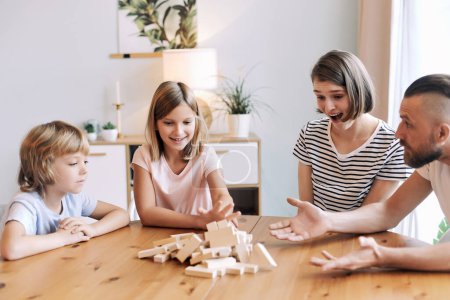 Photo for Loving young parents with little son and daughter enjoy playtime on leisure, play blocks removal game seated at table in living room on weekend pastime at home. Offspring growth and development - Royalty Free Image