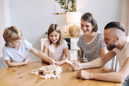 Photo for Loving young parents with little son and daughter enjoy playtime on leisure, play blocks removal game seated at table in living room on weekend pastime at home. Offspring growth and development - Royalty Free Image