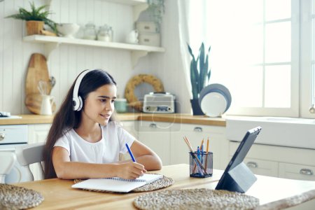 Téléchargez les photos : Jolie pré-adolescente 12s fille dans les écouteurs sans fil assis à la table e-learning, écouter des cours en ligne, leçon audio, recevoir de nouvelles connaissances, compétences en utilisant Internet et la technologie moderne. Développement de l'enfant. Éducation - en image libre de droit