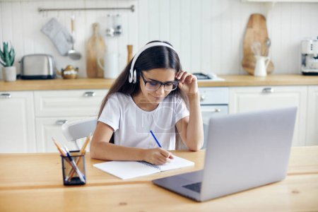 Photo for Pre-teen 12s girl in headphones and eyeglasses sit at table e-learns, listen online course, audio lesson, get new knowledge, skills using internet and modern tech. Development, education, eye-sight - Royalty Free Image