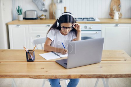 Photo for Pre-teen 12s girl in headphones and eyeglasses sit at table e-learns, listen online course, audio lesson, get new knowledge, skills using internet and modern tech. Development, education, eye-sight - Royalty Free Image
