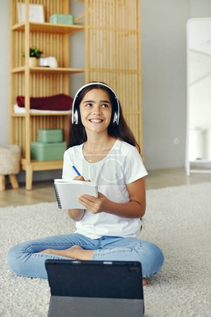 Photo for Pretty pre-teen girl sit on floor in cozy living room wear wireless headphones studying online use digital tablet device. Self-education, younger gen and modern tech usage for learning, gain new skill - Royalty Free Image