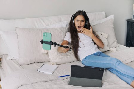 Photo for Attractive pre-teen girl resting seated on bed in bedroom holding selfie stick make photos, record video for social media channel, take part in live stream spend free time on internet. Vlogging, fun - Royalty Free Image