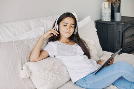 Photo for Cute pre-teen girl resting alone in bedroom with digital tablet device, wear wireless headphones enjoy favourite track, listen music on weekend at home. Young generation and tech usage, fun, leisure - Royalty Free Image