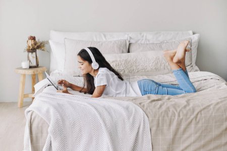 Photo for Adorable pre-teen girl in wireless headphones lying on bed with digital tablet device. Young generation and modern tech overuse, gadget usage for fun, hobby and leisure on weekend at home - Royalty Free Image